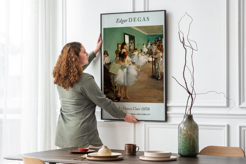 Pu&ograve; includere: Una donna appende una stampa d'arte incorniciata di "La Classe di Danza" (1874) di Edgar Degas su una parete bianca. La stampa presenta un gruppo di ballerine in uno studio di danza, con una parete verde chiaro e un pavimento in legno.