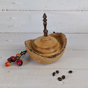 Könnte beinhalten: Eine handgefertigte Holzschale mit Deckel. Die Schale ist hellbraun mit einem dunkelbraunen Rand und einer dunkelbraunen Spitze auf dem Deckel. Sie steht auf einer weißen Holzoberfläche. Ein Zweig mit roten und orangen Beeren liegt neben der Schale.