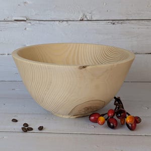 Op de afbeelding: Een lichtgekleurde houten kom met een brede, afgeronde vorm. De kom heeft zichtbare houtnerfpatronen en een gladde afwerking. Een takje met rode en oranje bessen en een paar koffiebonen liggen naast de kom.