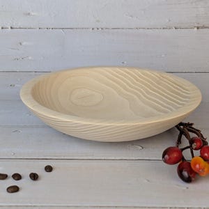 Op de afbeelding: Een lichtgekleurde houten kom met een gesneden, golvend interieur. De kom is rond met een ondiepe diepte. Een takje rode en oranje bessen en een paar koffiebonen liggen naast de kom op een wit houten oppervlak.
