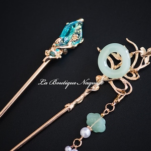 Può includere: Due bastoncini per capelli dorati con accenti decorativi di pietre preziose blu e verdi. La parte superiore di un bastoncino presenta una pietra preziosa blu con accenti floreali dorati. L'altro bastoncino ha un anello di giada verde con accenti floreali dorati e un ciondolo di giada verde e una perla.