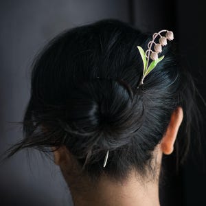 Peut inclure: Une épingle à cheveux décorative avec un motif de muguet. L'épingle a une tige argentée avec des feuilles vertes et de petites fleurs roses claires en forme de cloche. L'épingle à cheveux est placée dans un chignon.