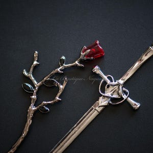 Può includere: Una spada d'argento con una rosa rossa e foglie nere su uno sfondo nero.