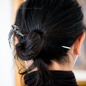 Può includere: Un fermaglio per capelli a forma di mano scheletrica argentata con uno spillo lungo e sottile argentato. Il fermaglio è nei capelli neri di una donna, che sono tirati indietro in uno chignon.