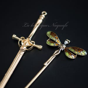 May include: Gold-toned metal hair accessories, including a sword-shaped pin and a dragonfly-shaped hair stick with green and brown enamel wings. The accessories are set against a black background.