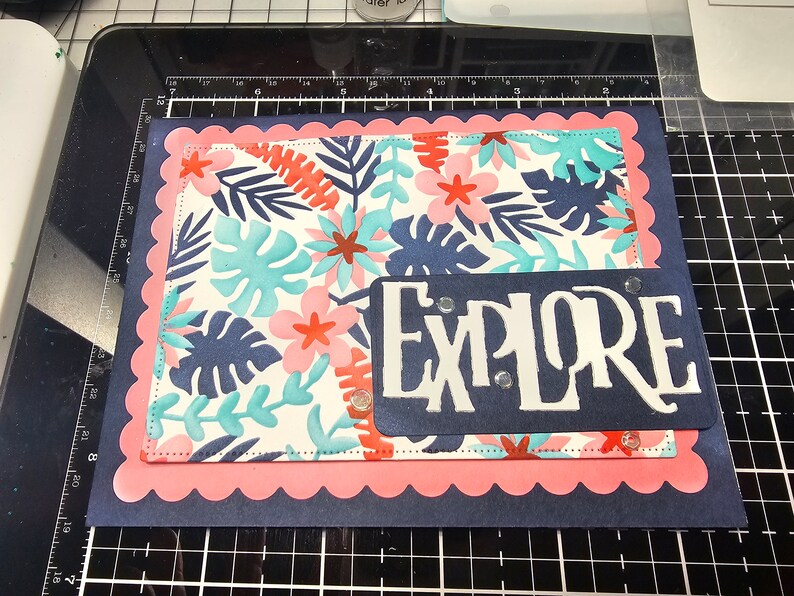 May include: A handmade greeting card with a tropical floral design. The card features a navy blue base, a pink scalloped border, and a patterned center with blue, pink, and coral flowers and leaves. The word "EXPLORE" is cut out in white on a navy blue rectangle.