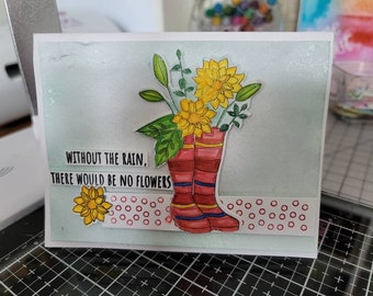 May include: A handmade greeting card featuring a watercolor illustration. Red rain boots hold yellow flowers and greenery. The text "WITHOUT THE RAIN, THERE WOULD BE NO FLOWERS" is printed on the card. A decorative strip with red circles accents the bottom.
