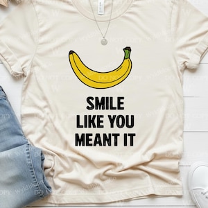 Puede incluir: Camiseta color crema con un gráfico de plátano amarillo y el texto "SMILE LIKE YOU MEANT IT" en negro. La camiseta se combina con vaqueros azules y zapatillas blancas. Se ve un collar plateado.
