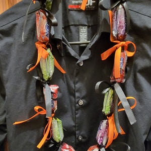 May include: A candy lei made of wrapped candy bars, tied together with black and orange ribbons. The lei is draped over a black button-down shirt hanging on a hanger. The candy bars are various colors and brands.