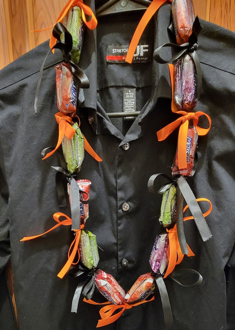 May include: A candy lei made of wrapped candy bars, tied together with black and orange ribbons. The lei is draped over a black button-down shirt hanging on a hanger. The candy bars are various colors and brands.