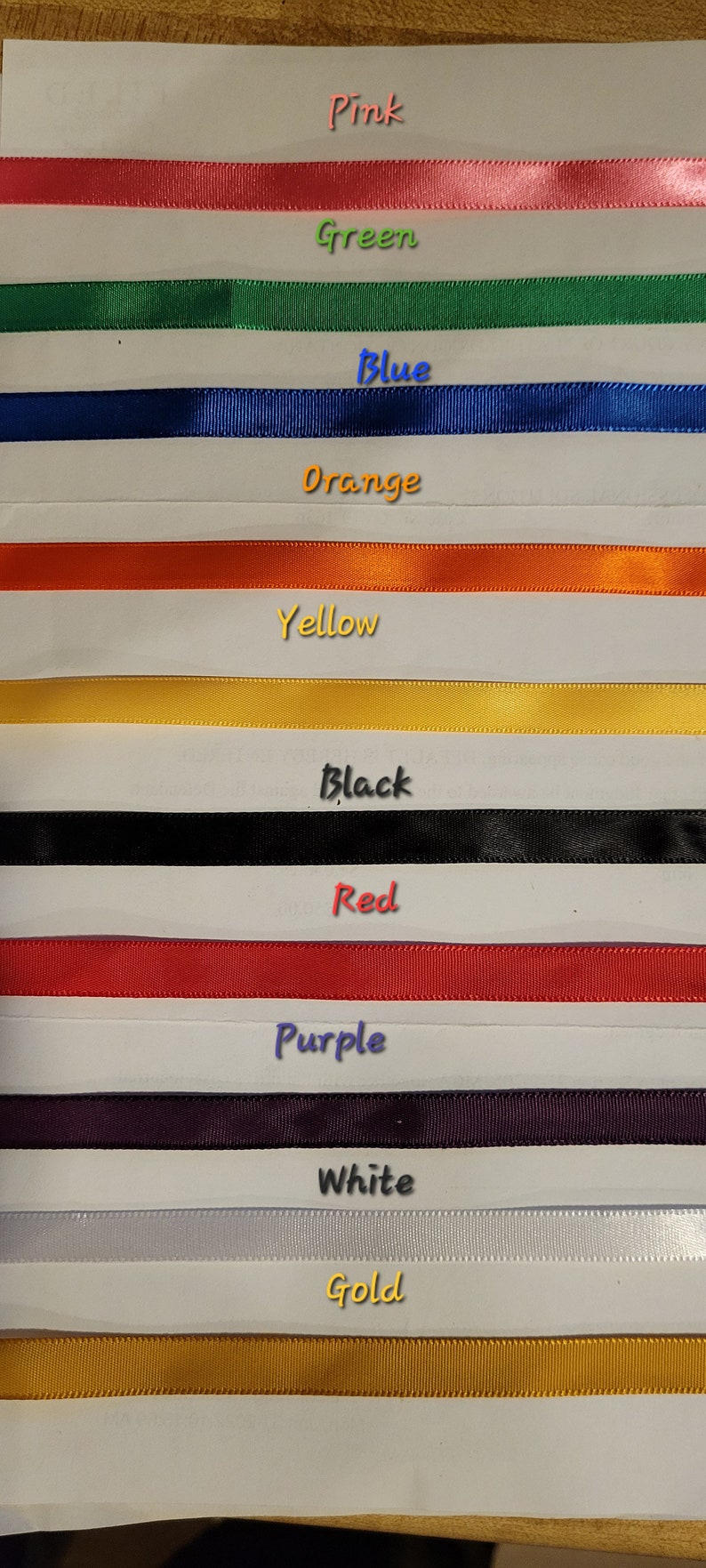 May include: A selection of colorful ribbons in pink, green, blue, orange, yellow, black, red, purple, white, and gold. Each ribbon is displayed horizontally with the color name written above it.