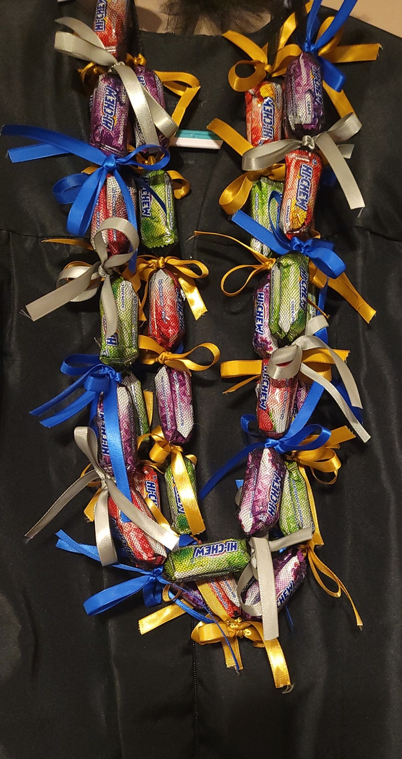 May include: A candy lei made of individually wrapped Hi-Chew candies in various colors, tied together with colorful ribbons. The ribbons are in shades of blue, gold, and silver. The lei is displayed against a black background.