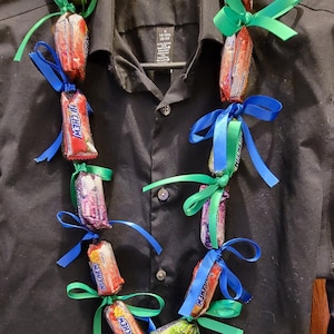 May include: A candy lei made of individually wrapped Hi-Chew candies, alternating with blue and green ribbons. The lei is displayed on a black button-down shirt.