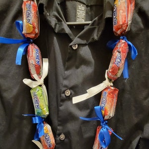 May include: A candy lei made of individually wrapped Hi-Chew candies in various colors, tied together with white and blue ribbons. The lei is displayed against a black button-down shirt.