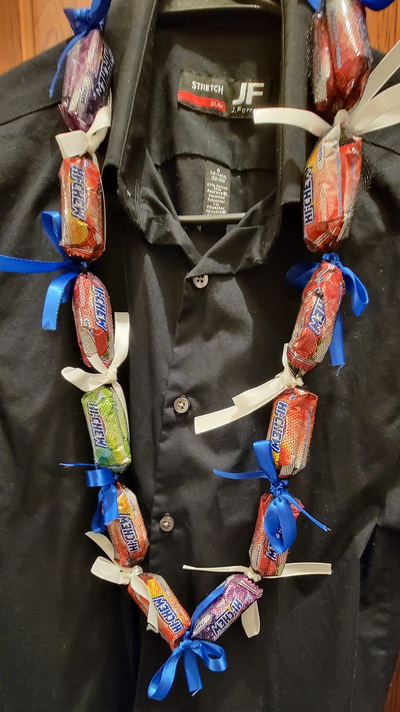 May include: A candy lei made of individually wrapped Hi-Chew candies in various colors, tied together with white and blue ribbons. The lei is displayed against a black button-down shirt.