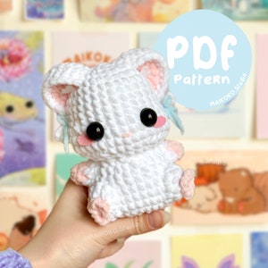 Op de afbeelding: Een wit gehaakt amigurumi-speelgoedkonijn met roze accenten en zwarte ogen. Het konijn wordt vastgehouden door een hand. De afbeelding bevat de tekst "PDF Pattern" in een blauwe cirkel.