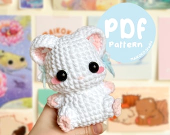 May include: A white crocheted bunny amigurumi toy with pink accents and black eyes. The bunny is being held by a hand. The image includes the text "PDF Pattern" in a blue circle.
