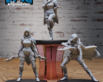 Puede incluir: Tres figuras en miniatura pintadas de gris que representan personajes femeninos con capas con capucha y sosteniendo dagas. Las figuras están en diferentes posturas y están sobre bases negras. Las figuras están sobre un fondo de pared de ladrillo.