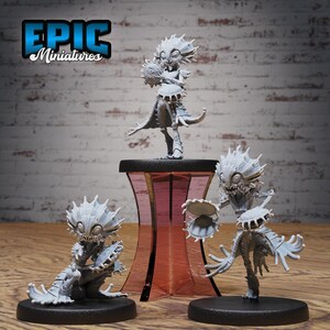 May include: Three gray, detailed, miniature figurines of creatures with large eyes and spiky hair playing musical instruments. The figurines are on black bases and a pink pedestal.