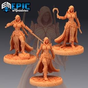 May include: Three miniature figures of female elves wearing hooded cloaks and armor, holding weapons. The figures are posed in different stances and are standing on circular bases.