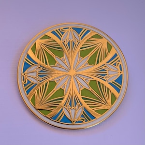 K&ouml;nnte beinhalten: Eine runde, goldfarbene Anstecknadel mit einem geometrischen Design in Wei&szlig;, Gr&uuml;n und Blau. Das Design zeigt stilisierte Blumen und Bl&auml;tter.
