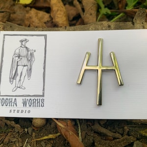May include: A silver metal pin with a stylized cross design. The pin is on a white card with the text "Pooha Works Studio".