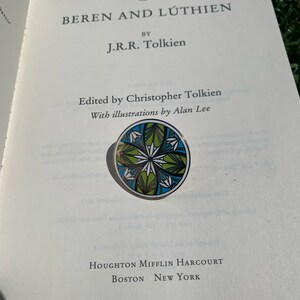 K&ouml;nnte beinhalten: Ein Buch mit dem Titel "Beren und L&uacute;thien" von J.R.R. Tolkien. Der Buchumschlag ist wei&szlig; mit schwarzem Text. Das Buch ist auf einer Seite ge&ouml;ffnet, auf der der Text "Herausgegeben von Christopher Tolkien" und "Mit Illustrationen von Alan Lee" steht. Eine runde, gr&uuml;ne, blaue und wei&szlig;e Emaille-Anstecknadel mit einem floralen Design befindet sich auf dem ge&ouml;ffneten Buch.