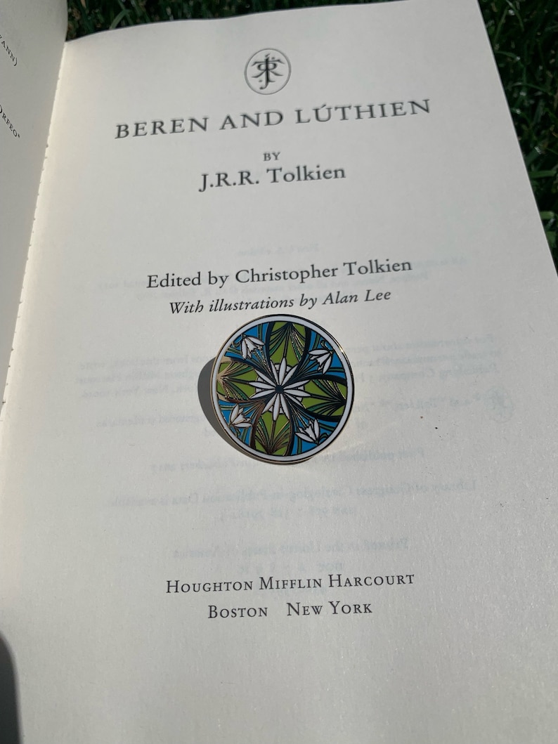 K&ouml;nnte beinhalten: Ein Buch mit dem Titel "Beren und L&uacute;thien" von J.R.R. Tolkien. Der Buchumschlag ist wei&szlig; mit schwarzem Text. Das Buch ist auf einer Seite ge&ouml;ffnet, auf der der Text "Herausgegeben von Christopher Tolkien" und "Mit Illustrationen von Alan Lee" steht. Eine runde, gr&uuml;ne, blaue und wei&szlig;e Emaille-Anstecknadel mit einem floralen Design befindet sich auf dem ge&ouml;ffneten Buch.