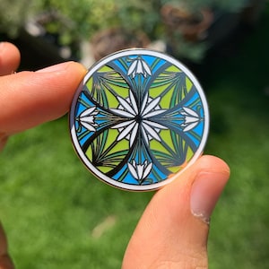 K&ouml;nnte beinhalten: Eine runde Emaille-Anstecknadel mit einem geometrischen Design, das stilisierte Blumen in Gr&uuml;n-, Blau- und Wei&szlig;t&ouml;nen zeigt.