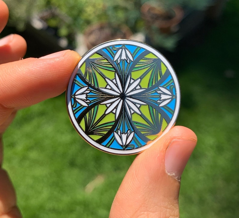 K&ouml;nnte beinhalten: Eine runde Emaille-Anstecknadel mit einem geometrischen Design, das stilisierte Blumen in Gr&uuml;n-, Blau- und Wei&szlig;t&ouml;nen zeigt.