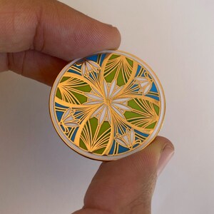 K&ouml;nnte beinhalten: Eine goldfarbene Emaille-Anstecknadel mit einem geometrischen Design in Wei&szlig;, Gr&uuml;n und Blau. Das Design zeigt eine stilisierte Blume mit Bl&uuml;tenbl&auml;ttern und Bl&auml;ttern.