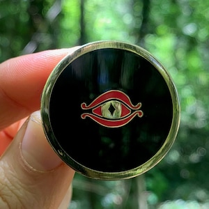 Puede incluir: Un pin de color dorado con fondo negro y un ojo rojo con un diamante en el centro. El ojo está rodeado por un diseño dorado.
