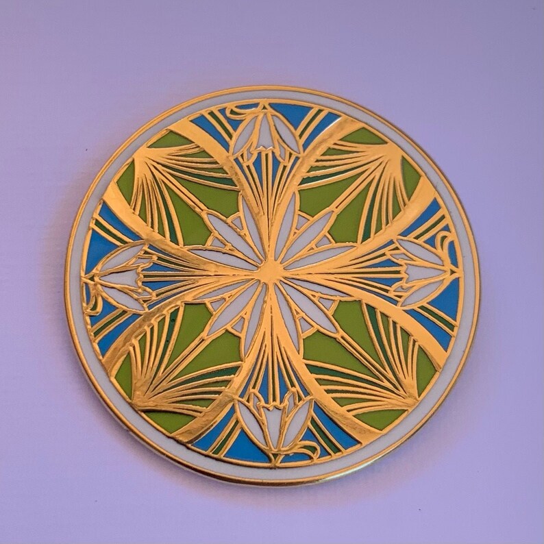 K&ouml;nnte beinhalten: Eine runde, goldfarbene Anstecknadel mit einem geometrischen Design in Wei&szlig;, Gr&uuml;n und Blau. Das Design zeigt stilisierte Blumen und Bl&auml;tter.