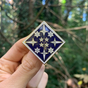 Puede incluir: Un pin de esmalte dorado con forma de rombo azul. El pin presenta un diseño de estrella blanca con un círculo blanco en el centro de cada estrella.