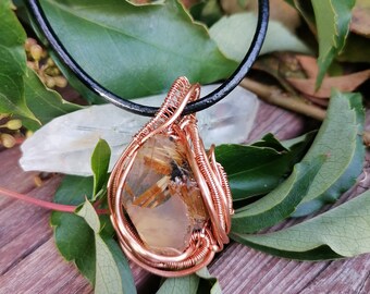 May include: A gold and brown crystal pendant wrapped in copper wire, hanging from a black leather cord. The pendant is surrounded by green leaves.