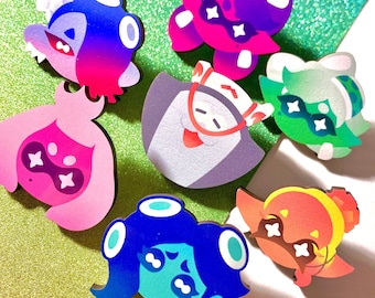 May include: A collection of colorful, cartoon-style character pins. The pins feature various designs with vibrant gradients of pink, purple, blue, green, and orange. Each pin has a unique face and shape, with some wearing glasses or hats.