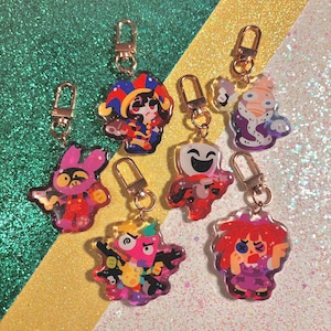 May include: A collection of colorful acrylic keychains with gold-colored clasps. The keychains feature various cartoon character designs in shades of red, pink, blue, and purple. The background is a glittery mix of green, gold, and white.