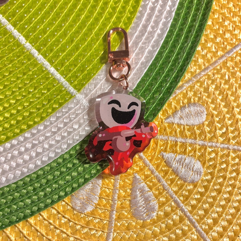 May include: A clear acrylic keychain featuring a cartoon character with a large, smiling face and a red instrument. The keychain has a gold-colored clasp and is set against a background of yellow, white, and green.