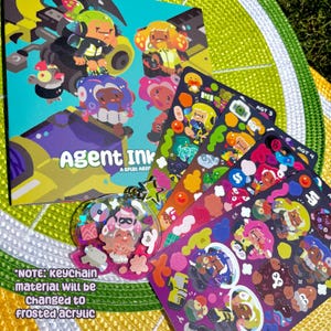 Puede incluir: Mercancía de Agent Ink que incluye un libro, hojas de pegatinas y un llavero. La portada del libro muestra personajes de dibujos animados en amarillo, verde y morado. Las hojas de pegatinas muestran varios diseños de personajes. El llavero incluye un dije de estrella. El texto "NOTE: keychain material will be changed to frosted acrylic" es visible.