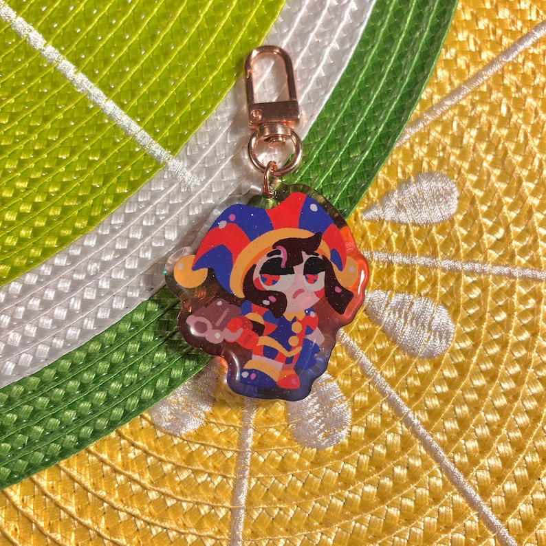 May include: A colorful acrylic charm featuring a cartoon jester character with a red, blue, and yellow hat. The charm has a gold-colored clasp. The background is a yellow and white woven pattern.