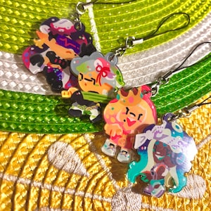 May include: Four colourful acrylic charms with cartoon character designs. Each charm is attached to a black lanyard with a silver-coloured clasp. The charms feature various colours, including pink, purple, green, and orange, against a yellow and green background.