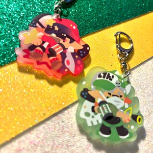 May include: Two acrylic keychains featuring colorful cartoon characters. One keychain has a red and black character, the other a green and white character. Both keychains have silver-colored clasps and are set against a glittery background.