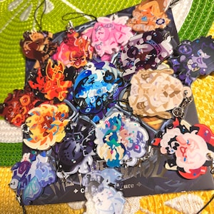 May include: A collection of colorful acrylic charms with various character designs. The charms feature a variety of colors, including red, blue, pink, and yellow, with attached keychains. The charms are arranged on a patterned surface.