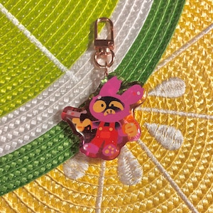 May include: A pink and red cartoon bunny keychain with a gold clasp. The bunny is wearing red overalls and holding a weapon. The background is a yellow and green woven pattern.