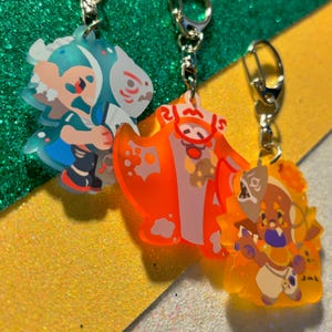 May include: Three colorful acrylic keychains with silver clasps. The keychains feature cartoon characters in shades of blue, orange, and yellow. The background is a mix of green and yellow glitter.