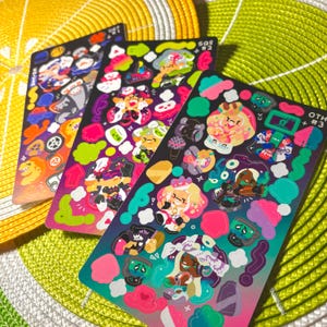 Puede incluir: Tres hojas de pegatinas coloridas con personajes de dibujos animados y varias formas. Las pegatinas presentan una variedad de colores, incluyendo rosa, verde, azul y amarillo. Las hojas están sobre una superficie con estampado verde y amarillo.