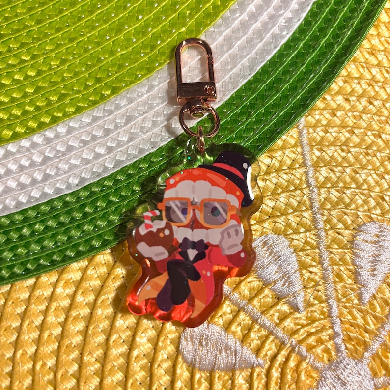 May include: A clear acrylic keychain featuring a cartoon Santa Claus character with orange glasses, a black top hat, and a red suit. The keychain has a gold-colored clasp and a small ring. The background is a mix of yellow, green, and white.