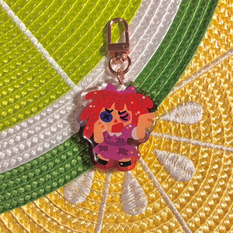 May include: A colorful acrylic keychain featuring a cartoon character with red hair, a purple dress, and a bow. The keychain has a gold-colored clasp and is set against a yellow and green background.