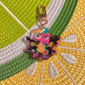 May include: A colorful acrylic keychain featuring a cartoon character with guns. The character is pink, yellow, and green, with black guns and a gold-colored clasp. The background is a lime and lemon-themed pattern.
