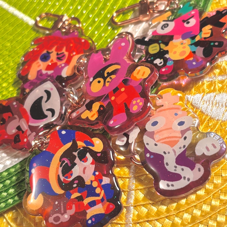 May include: A collection of colorful, cartoon-style acrylic keychains. Each keychain features a unique character design with vibrant colors and glitter accents. The keychains are attached to gold-colored clasps.